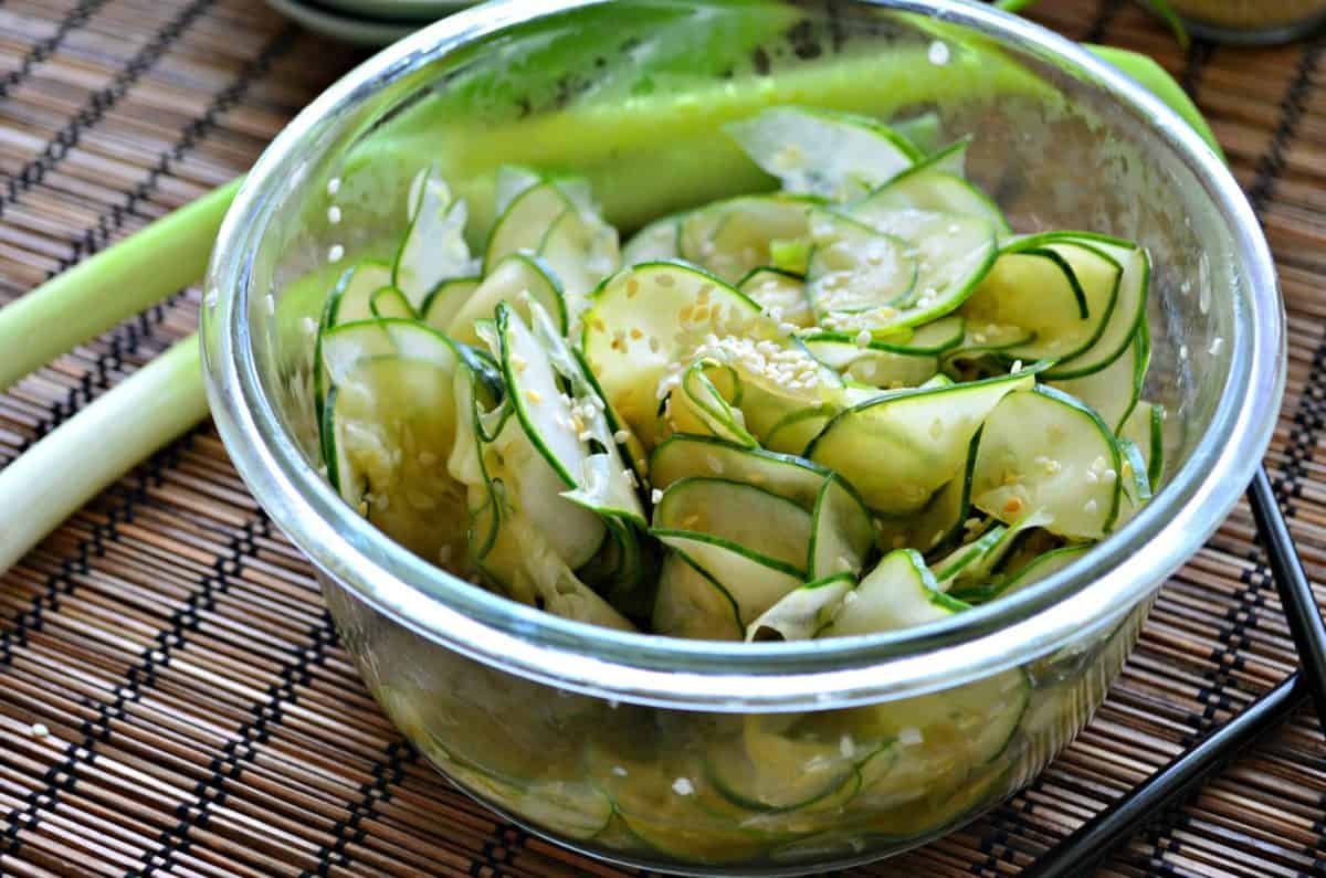 Glass bowl filled with thinly sliced cucumbers and sesame seeds on a wood mat. 