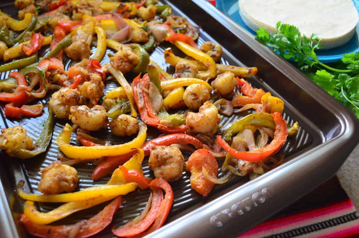 seasoned shrimp, sliced bell peppers, and onions on a sheet pan next to fresh cilantro and tortillas.