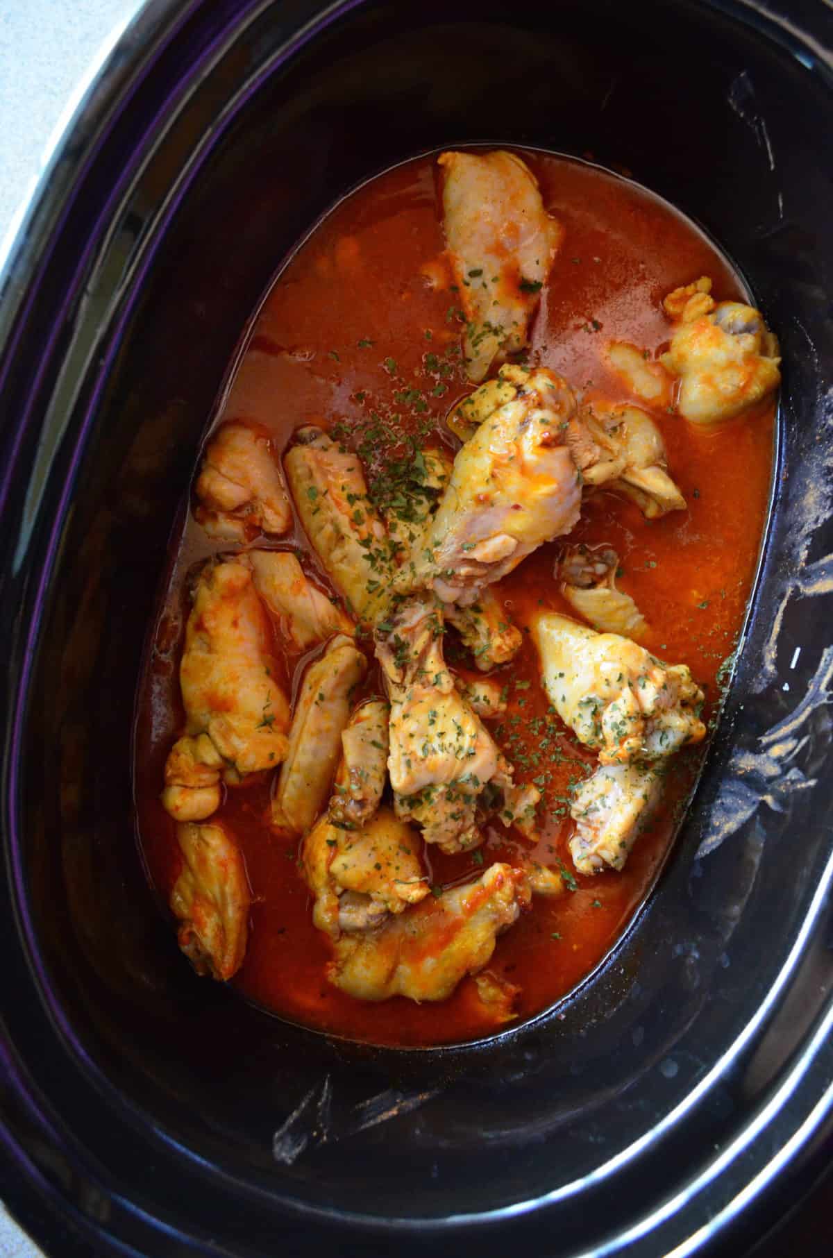 top view of chicken wings in buffalo sauce with herbs in slow cooker.