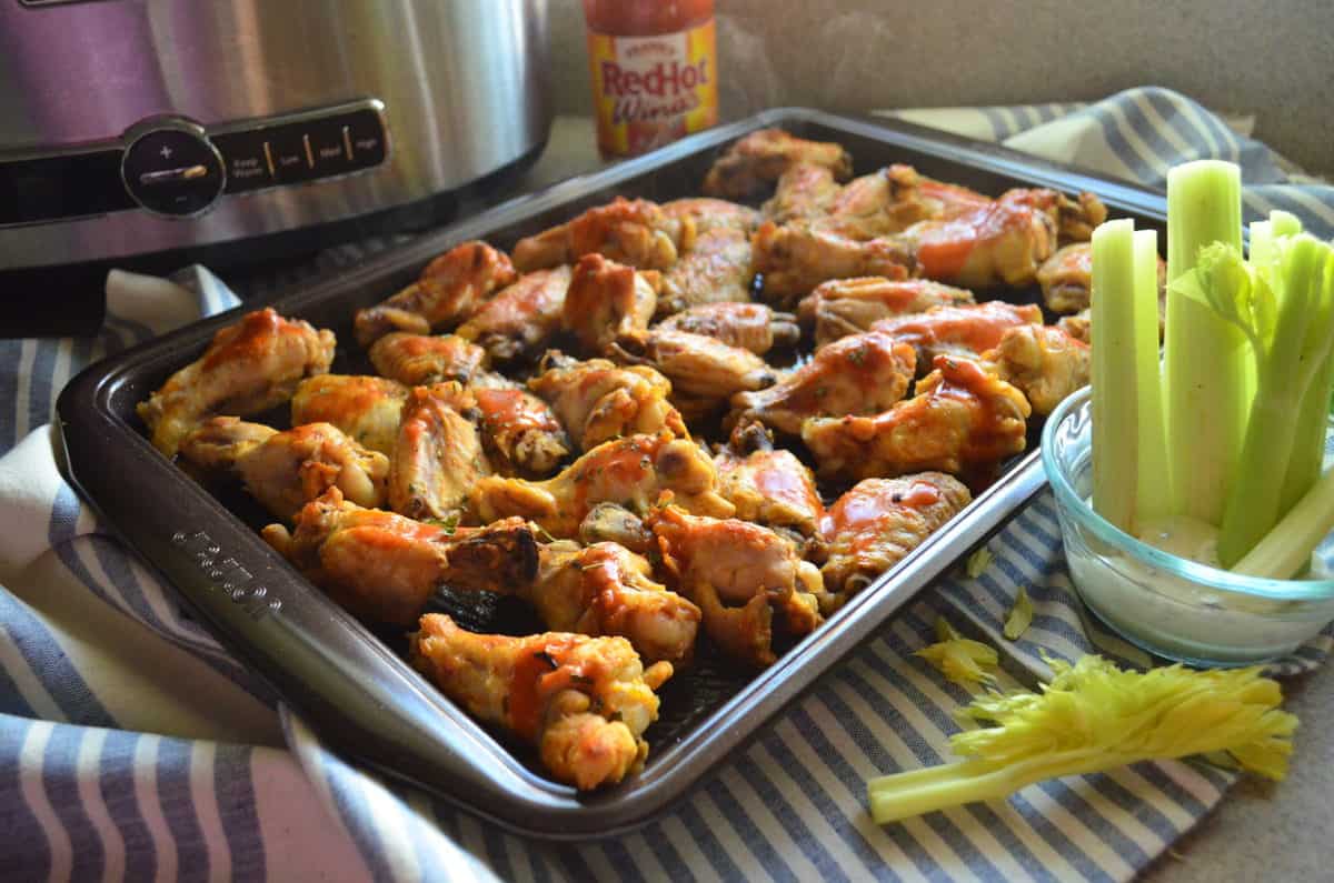 wings smothered in buffalo sauce and spices on sheet pan next to bowl of celery dipped in ranch dressing.