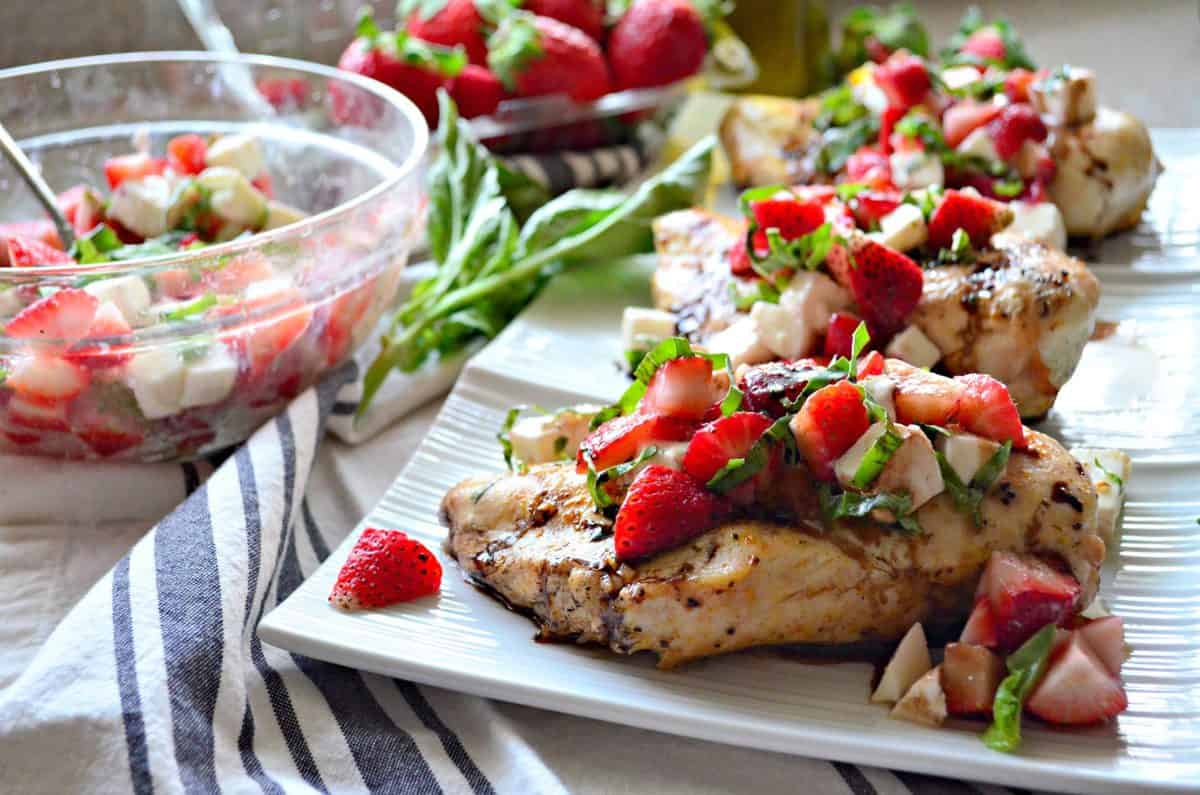 grilled chicken topped with basil, strawberries, and balsamic sauce on platter next to bowl of ingredients mixed.