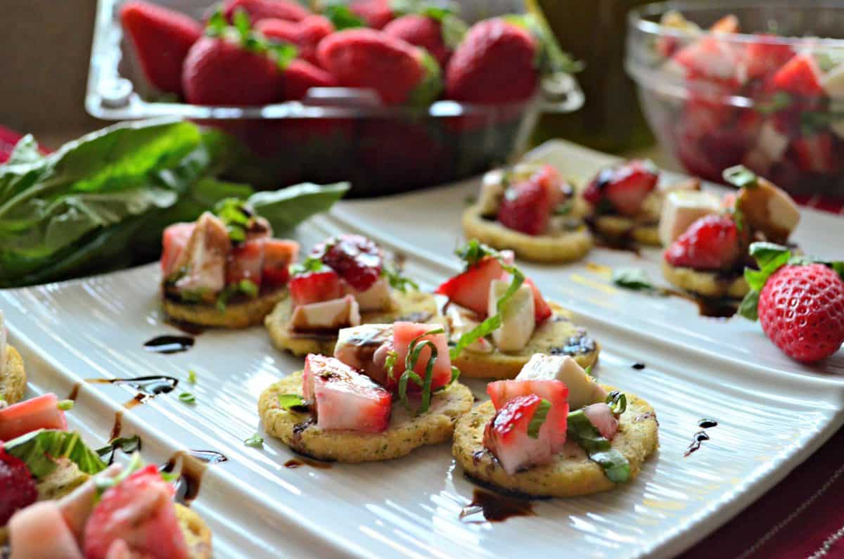 herbed crackers topped with strawberries, cheese, balsamic, and basil on white platter.