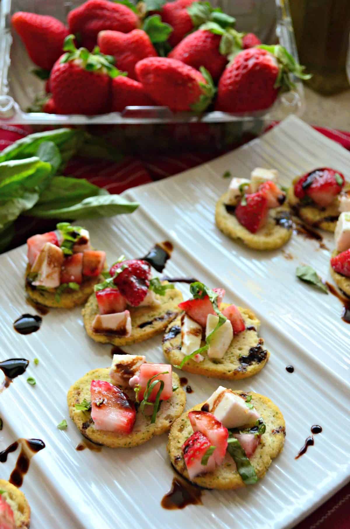 closeup of herbed crackers topped with strawberries, cheese, balsamic, and basil on white platter.