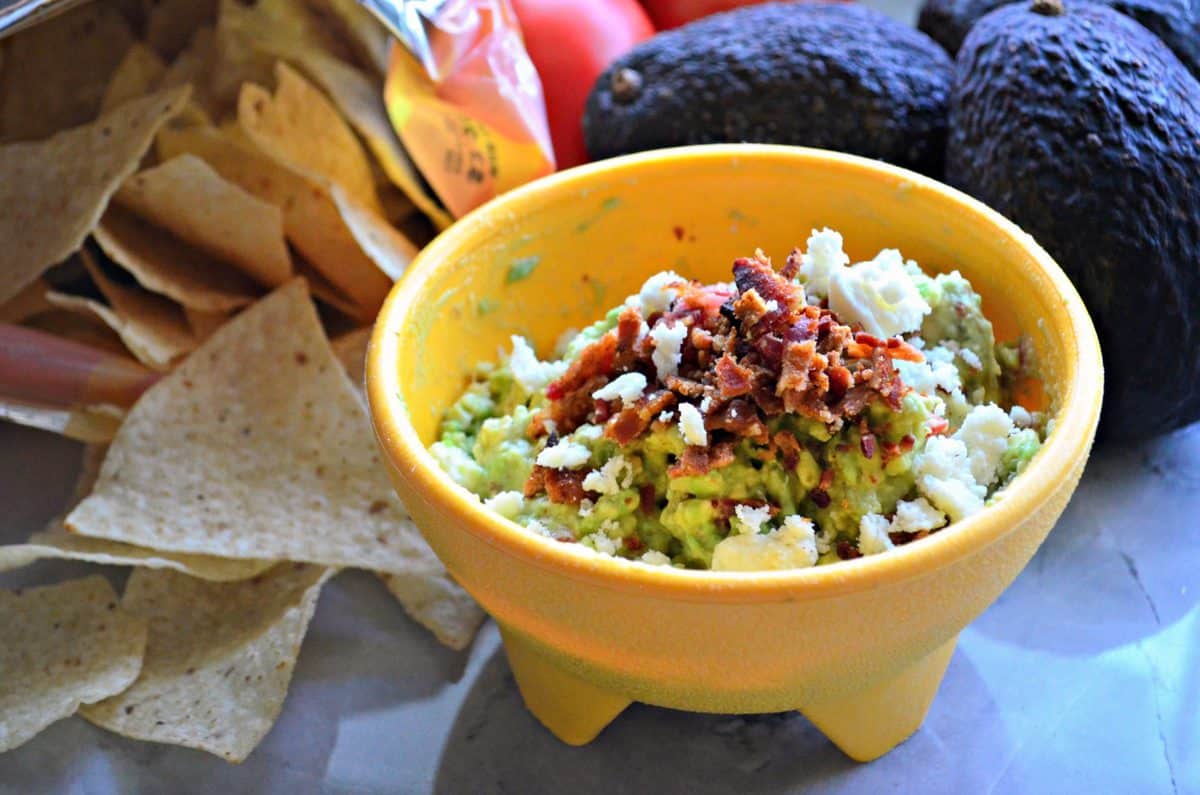 small yellow bowl of guacamole topped with queso fresco and bacon next to corn tortilla chips and avocados.