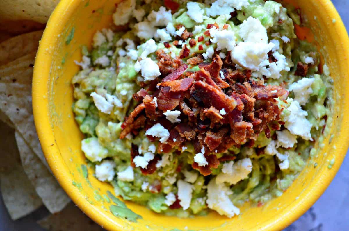close up top view of small yellow bowl of guacamole topped with queso fresco and bacon.