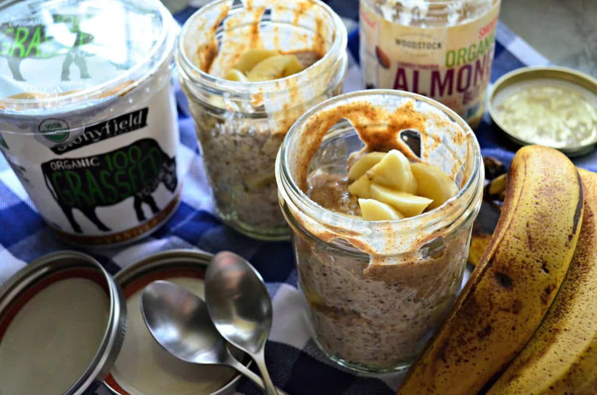 two mason jars of oats, bananas, and almond butter next to fresh bananas and spoons on tablecloth.