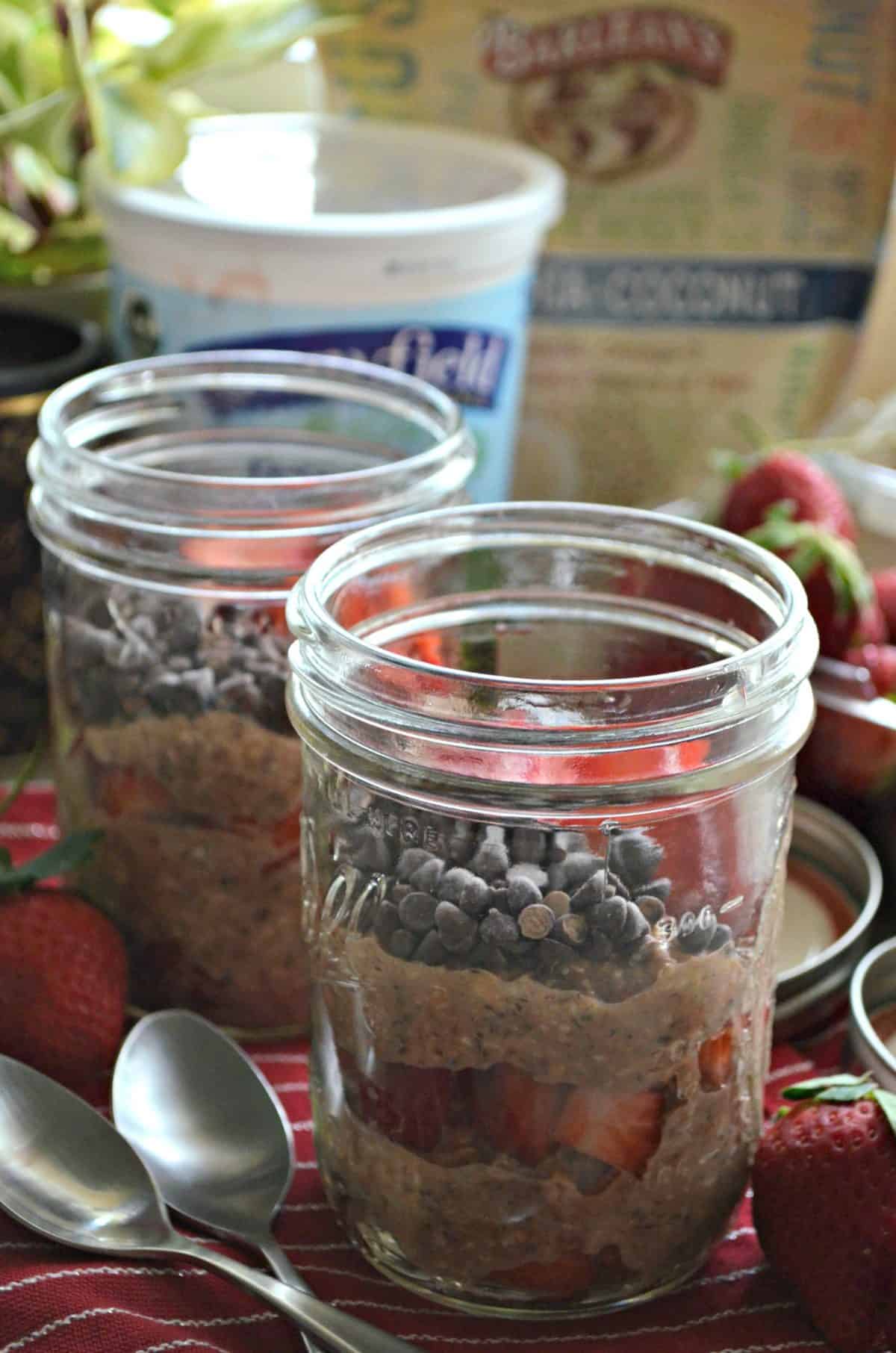 2 glass jars of layered oats, chocolate chips, and sliced strawberries with yogurt container in background.