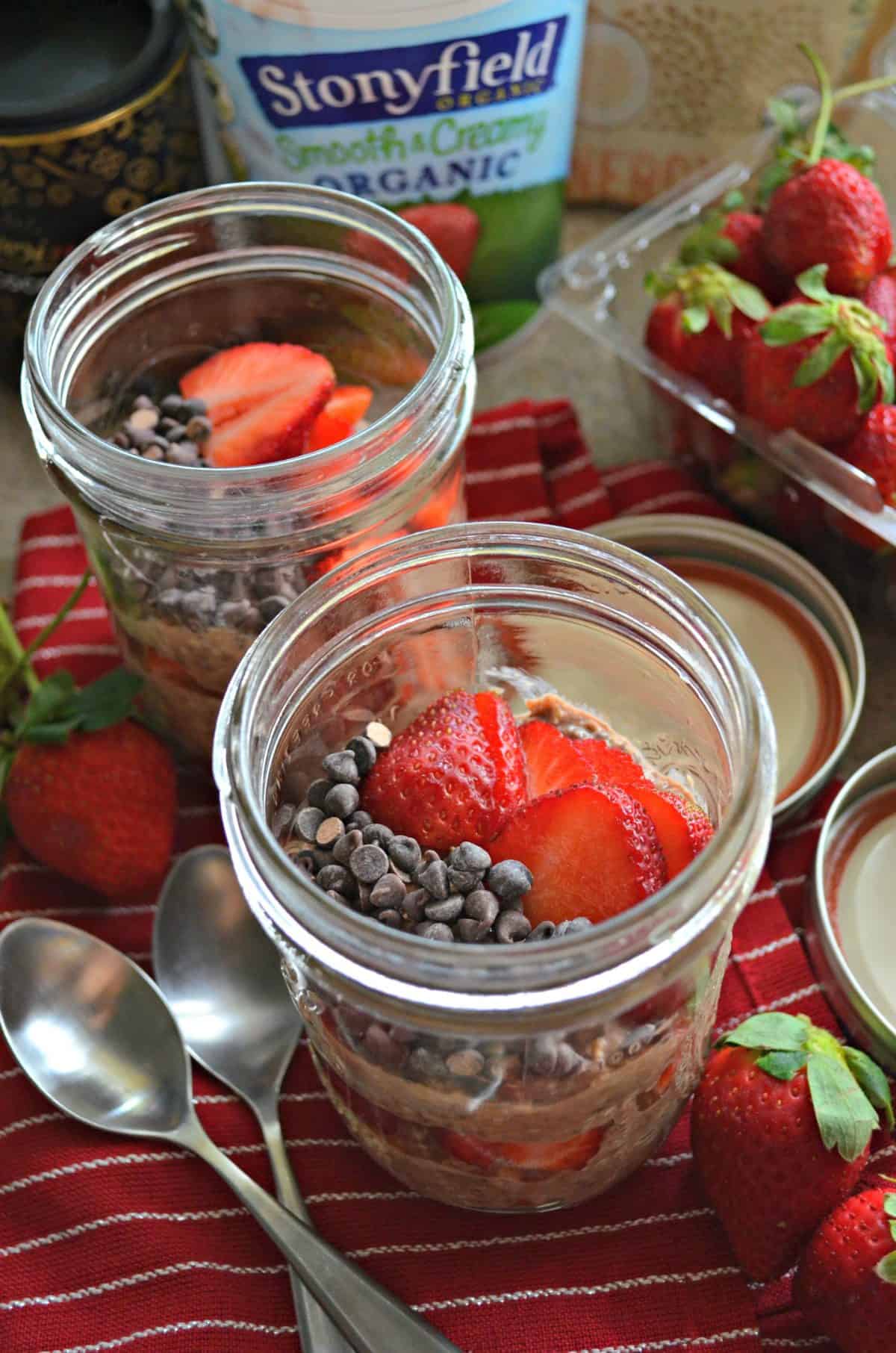 2 glass jars of layered oats, chocolate chips, and sliced strawberries on red tablecloth with strawberries and spoons.