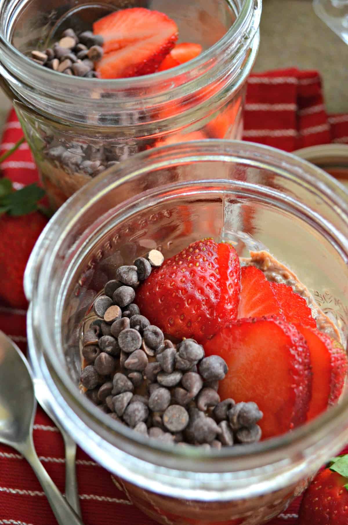 closeup 2 glass jars of layered oats, chocolate chips, and sliced strawberries.