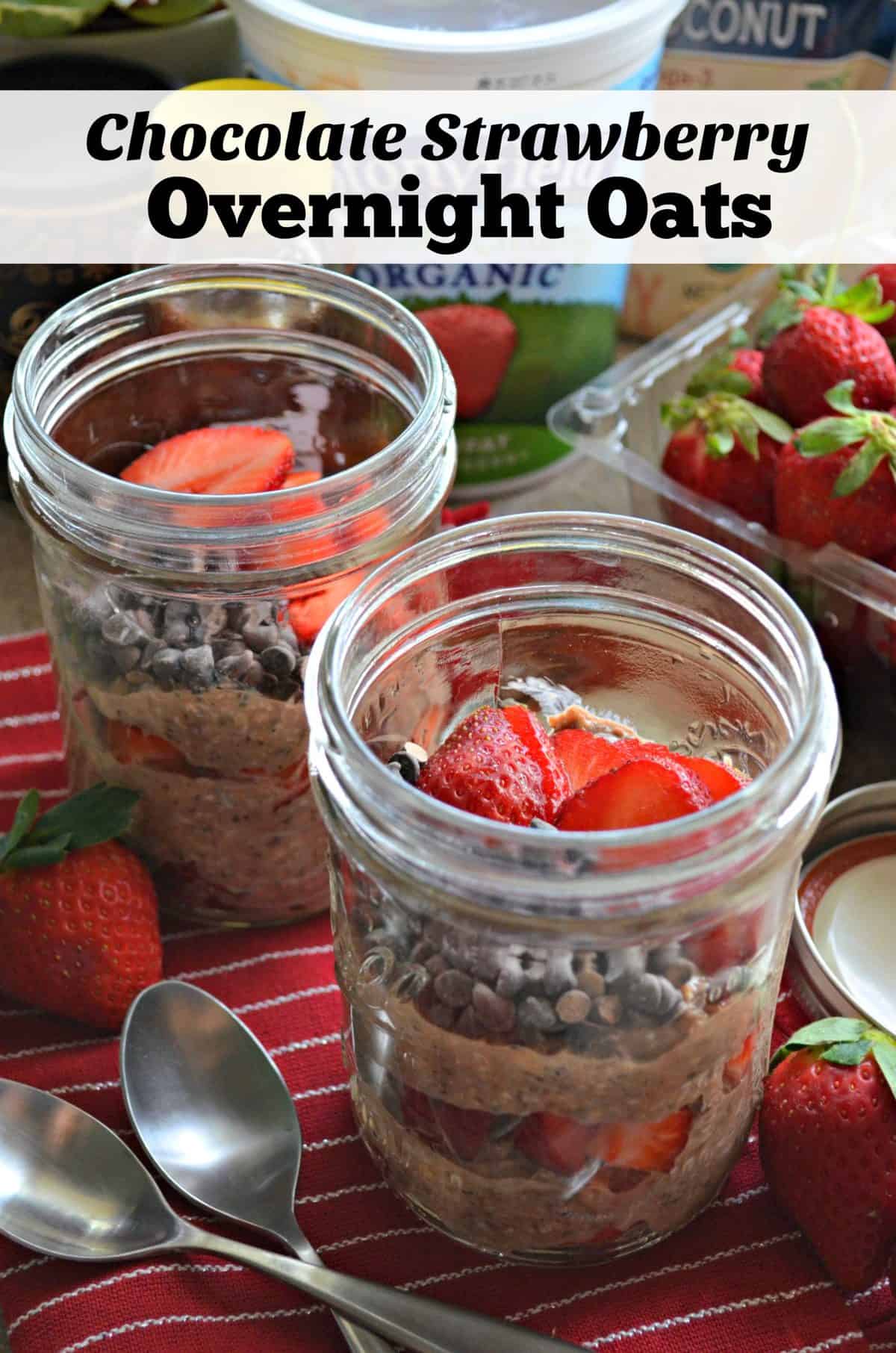 2 glass jars of layered oats, chocolate chips, and sliced strawberries on red tablecloth with strawberries.