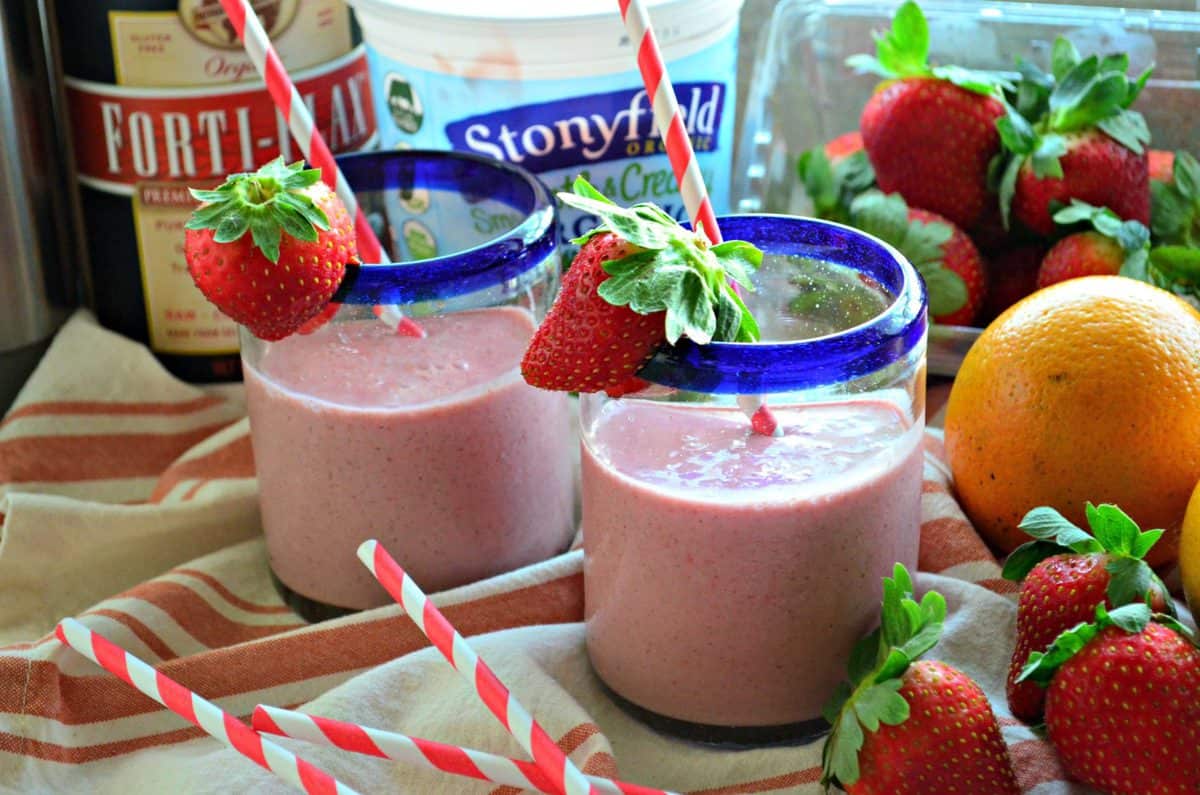 two blue rimmed glasses with red pink blended drink rimmed with fresh strawberries near fresh fruits.