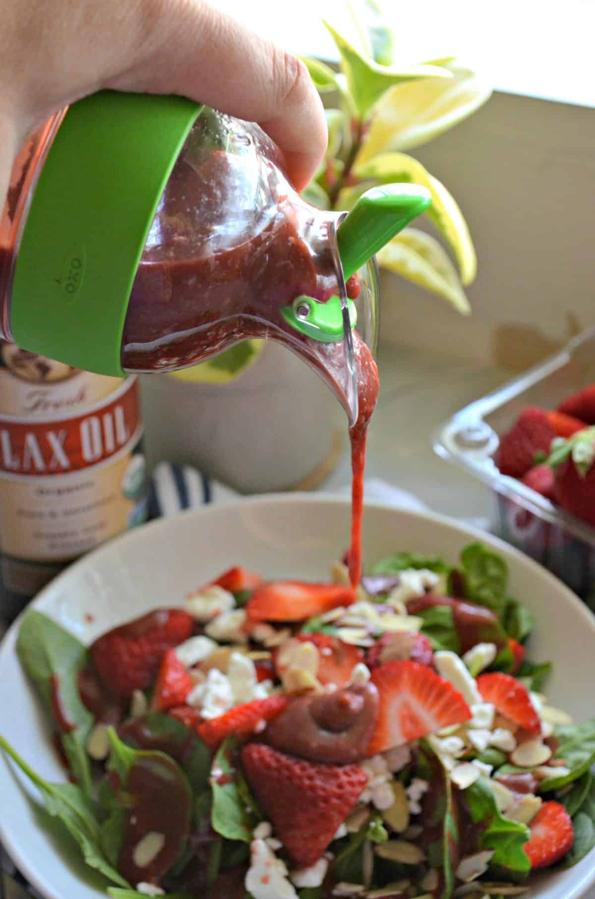 hand holding salad dressing container filled with pink seedy liquid over salad bowl as it drizzles down.