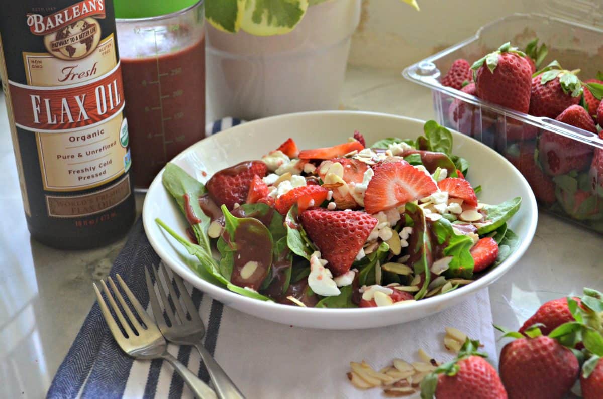 spinach salad with strawberries, almond slivers, dressing, and feta in white bowl next to barlean's flax oil.