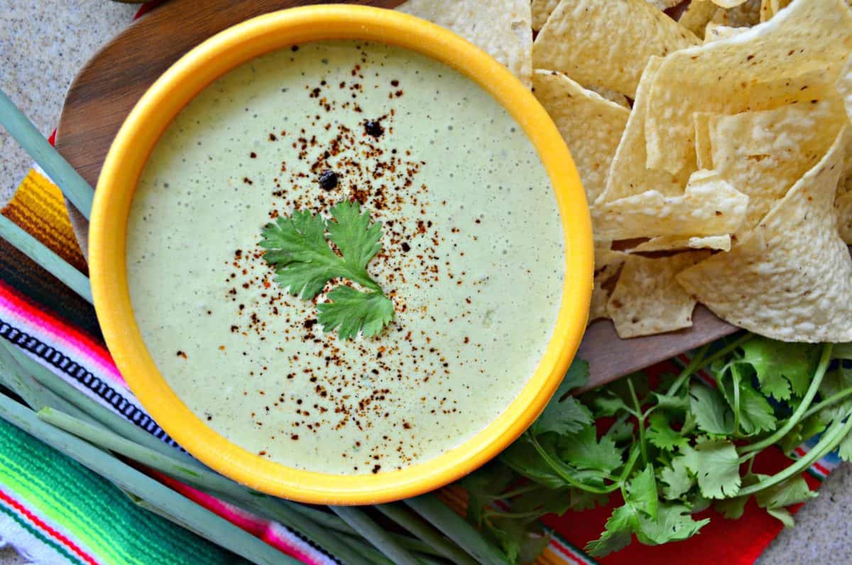 top view of creamy green dip in yellow bowl topped with cilantro leaf next to corn tortilla chips and cilantro.