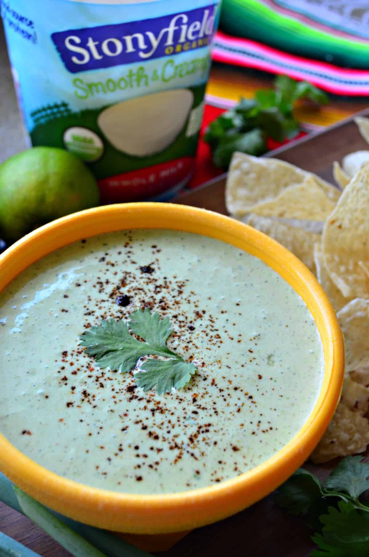 close up top view creamy green dip in yellow bowl topped with cilantro leaf next to corn tortilla chips and limes.