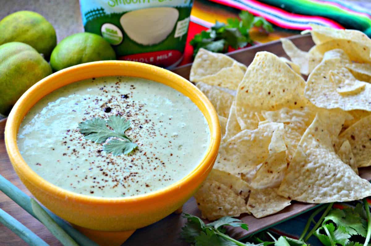 side view of creamy green dip in yellow bowl topped with cilantro leaf next to corn tortilla chips and cilantro.