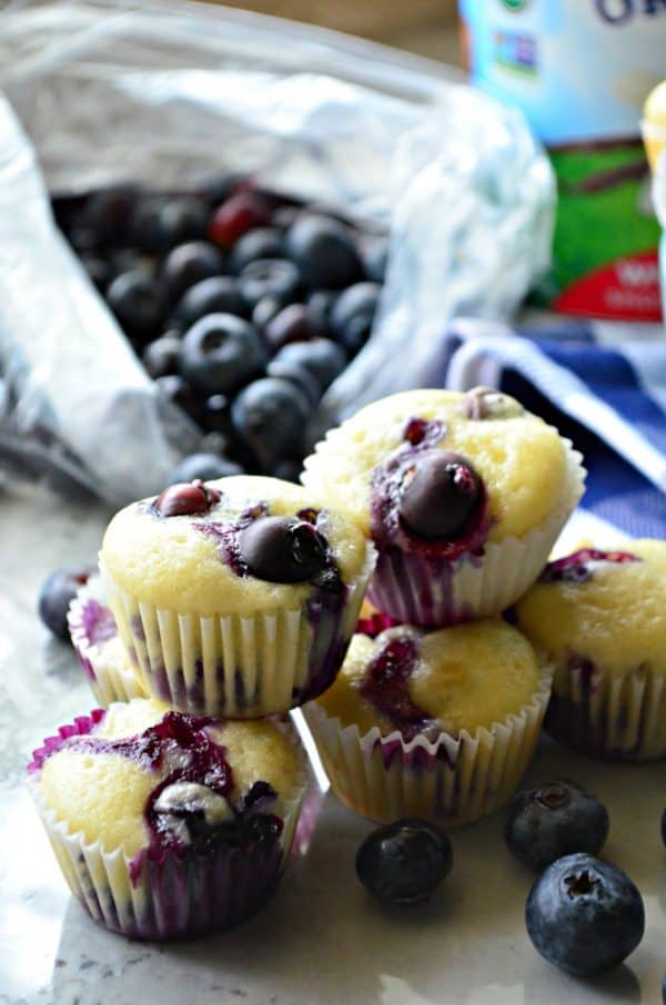 mini blueberry yogurt muffins stacked on countertop in front of bag of fresh blueberries.