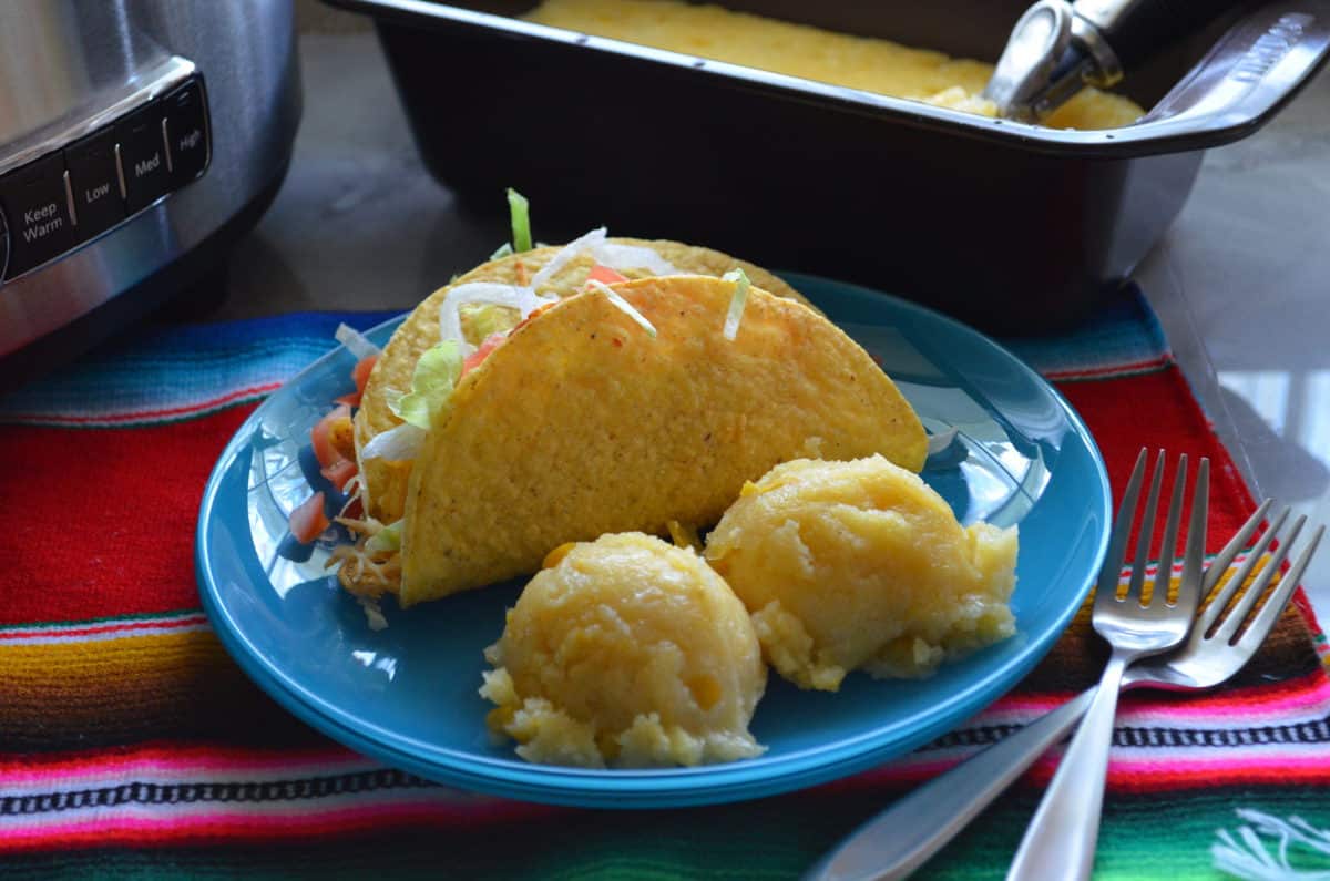 Hard shell taco with tomatoes and lettuce showing plated with two corn cakes on tablecloth with forks.