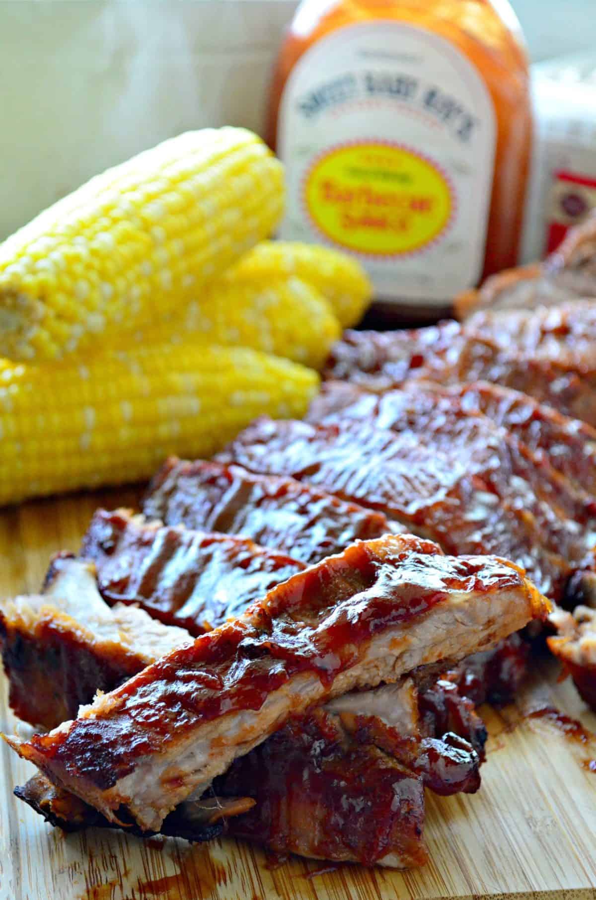 closeup baby back ribs smothered in red sauce and stacked on cutting board in front of fresh corn and BBQ sauce.