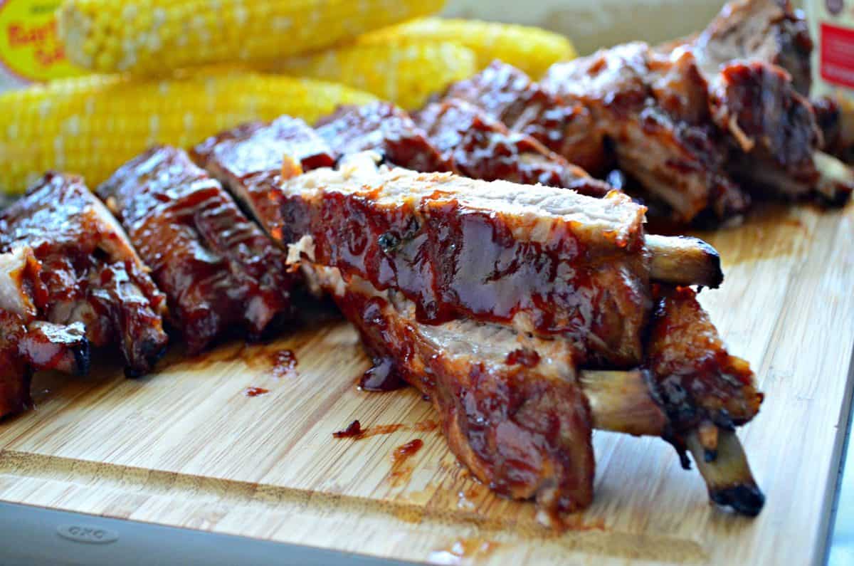 baby back ribs smothered in red sauce and stacked on cutting board in front of fresh corn.