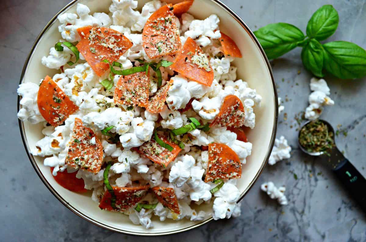 top view bowl of quartered pepperoni, basil, and seasonings on popcorn on countertop by fresh basil.