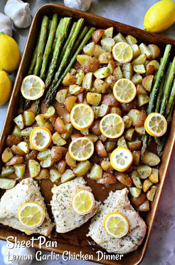 Top view of a brown sheet pan with diced ptoatoes, asparagus, and chicken breast with lemon wedges.