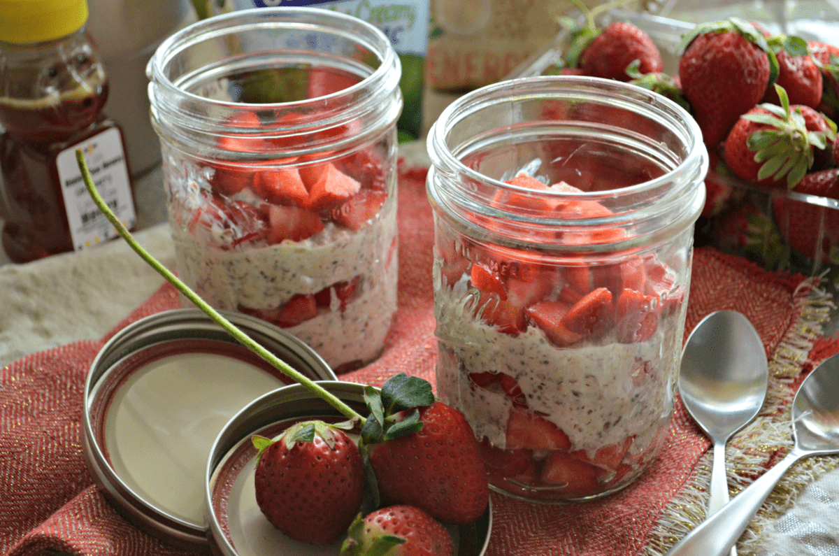 2 small mason jars filled with oatmeal looking substance and chopped strawberries with fresh strawberries in background.