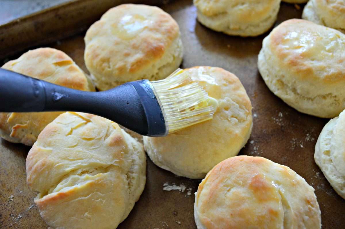 Silicone brush brushing butter on to a cooked biscuit on a baking tray.