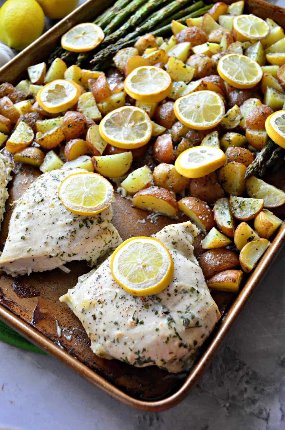 close up sheet pan of quartered punch potatoes, lemon wheels, asparagus, and chicken breast with herbs.