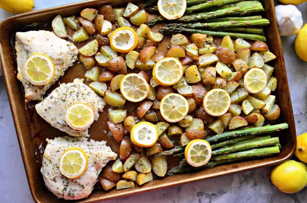 top view sheet pan of quartered punch potatoes, lemon wheels, asparagus, and chicken breast with herbs.