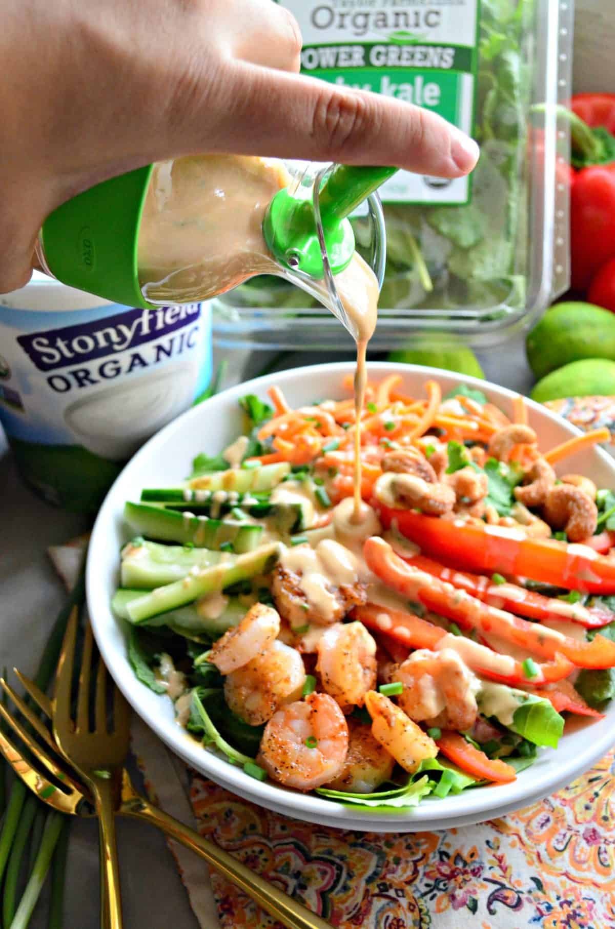 homemade dressing being poured over bowl of sliced cucumbers, kale, bell peppers, cashews, carrots, and seasoned shrimp.