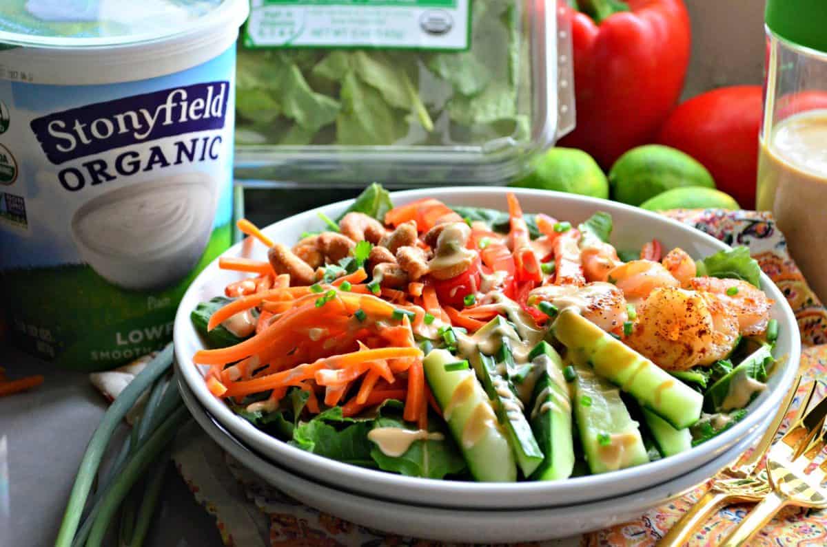 bowl of sliced cucumbers, kale, bell peppers, cashews, carrots, and seasoned shrimp with dressing in front of yogurt.