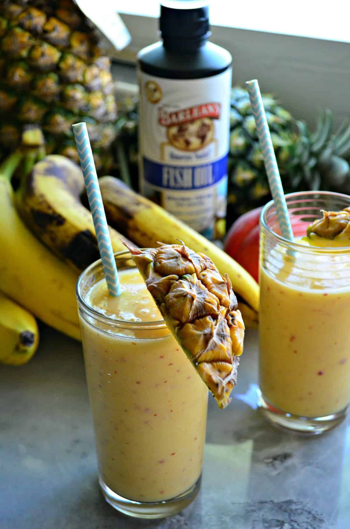 two tall glasses full of yellow smoothie with orange speckles rimmed with pineapple and a straw.