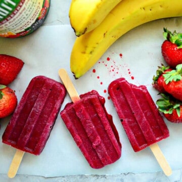 Three bright purple popsicles on white parchment paper with bananas and strawberries around it.