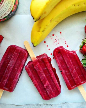 Three bright purple popsicles on white parchment paper with bananas and strawberries around it.