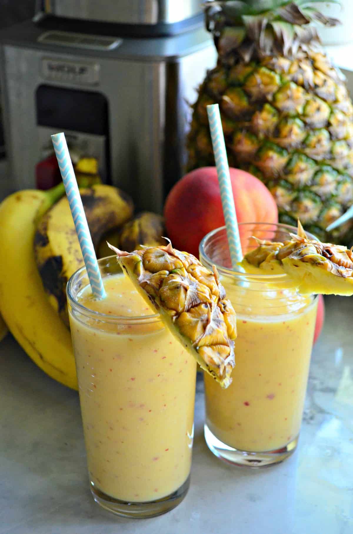 two tall glasses full of yellow smoothie rimmed with pineapple and a straws in front of fresh fruit.