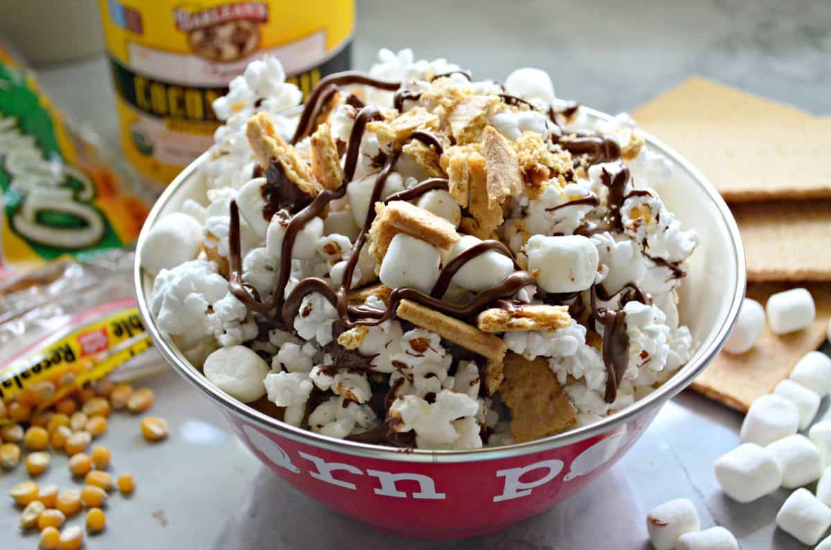 bowl of popcorn with graham crackers, chocolate drizzles, and mini marshmallows on countertop with ingredients blurred behind.