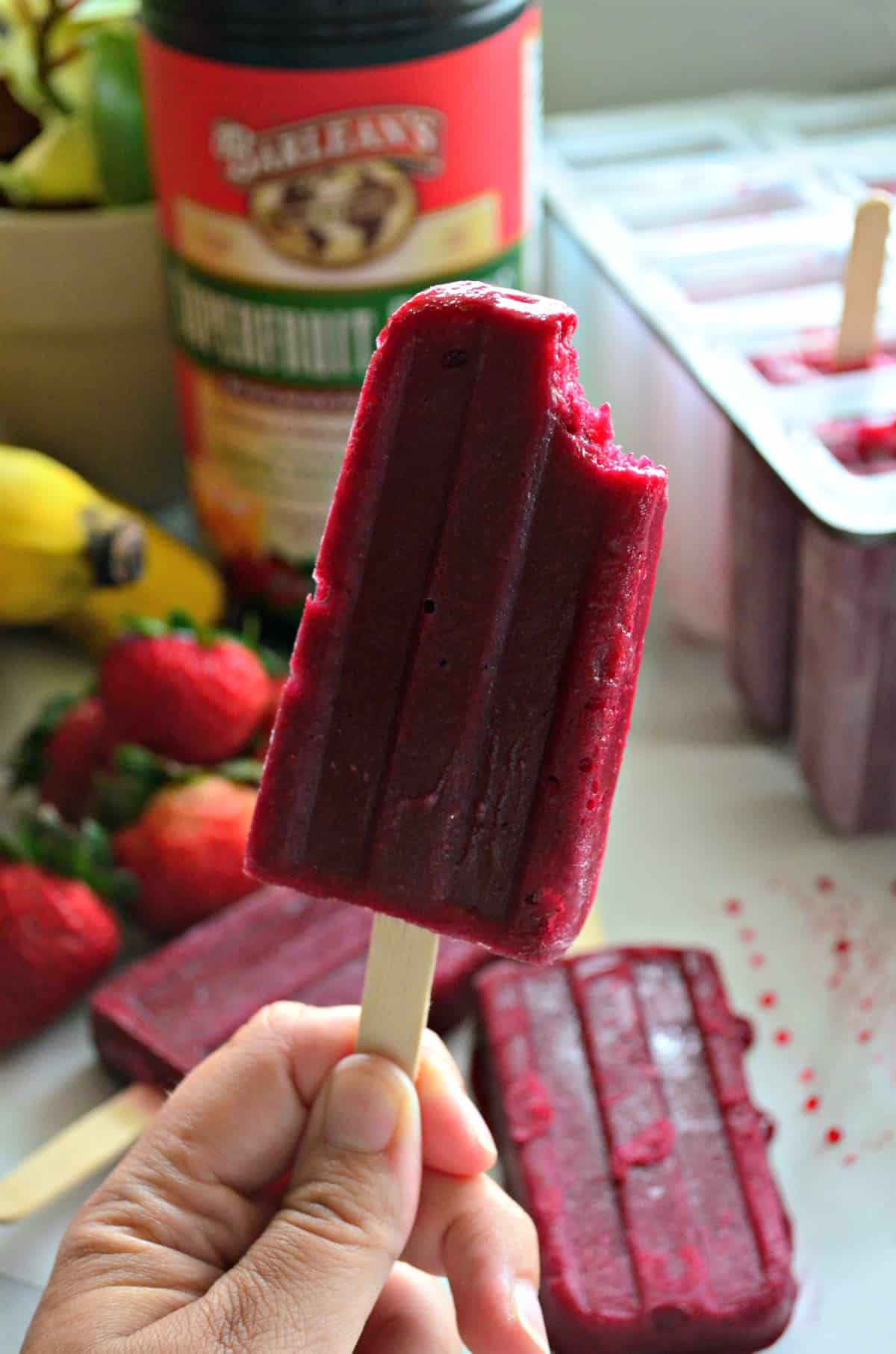 close up hand holding beet red popsicle with bite out of it and fruits blurred in background.