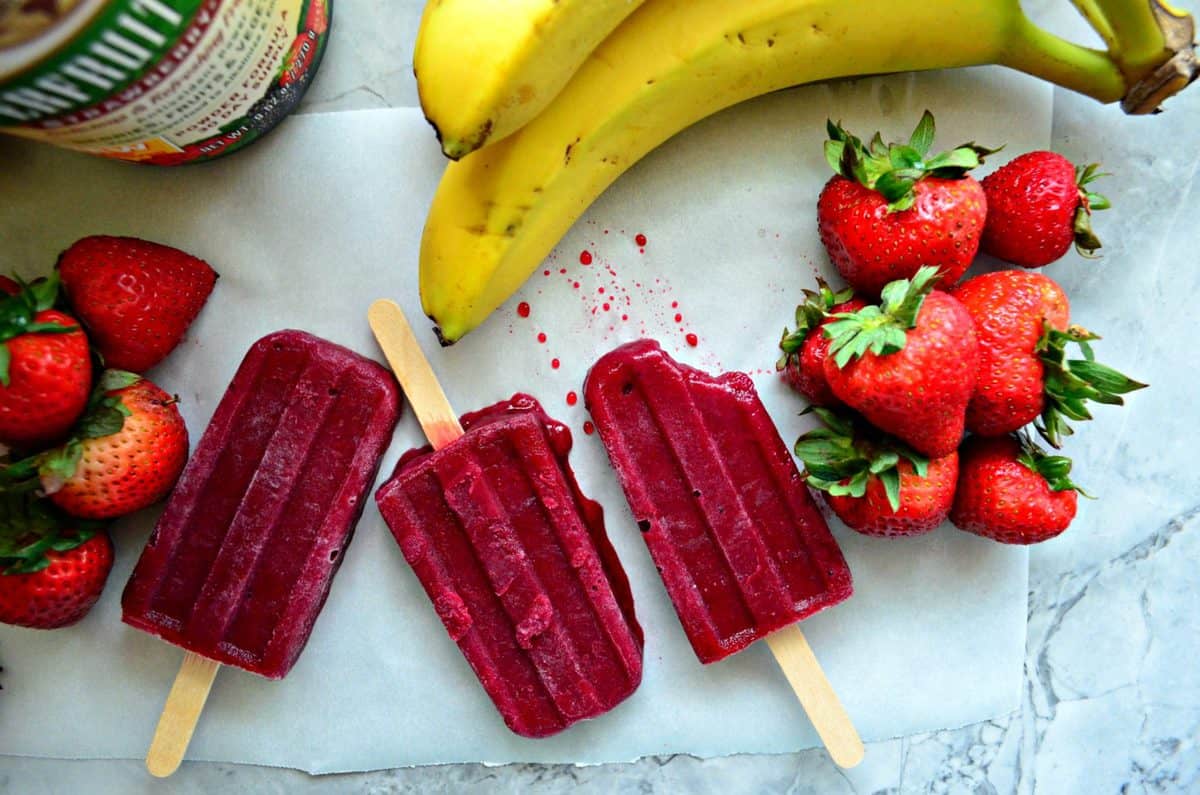 3 beet red popsicles side by side on parchment paper on countertop next to bananas and strawberries.