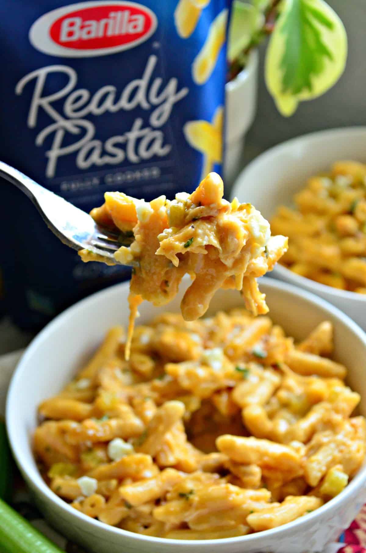 fork full of creamy light orange colored buffalo chicken penne pasta held over white bowl with herbs and cheese.