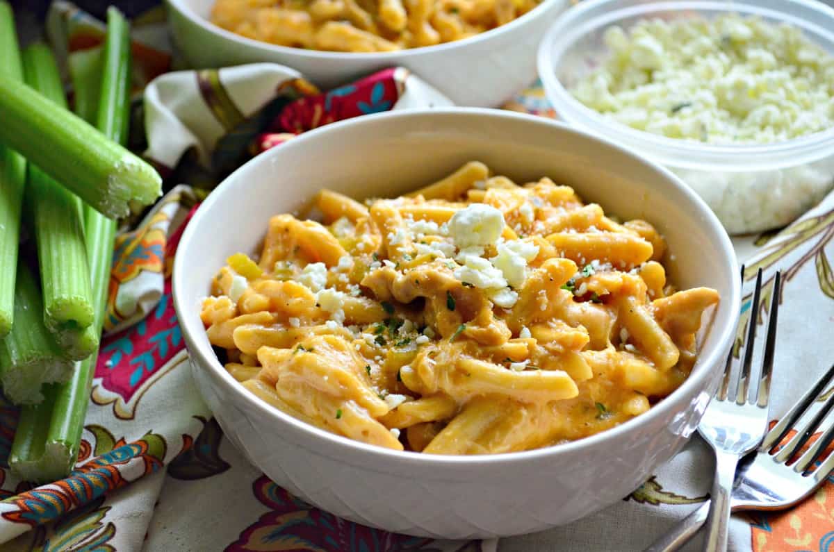 creamy light orange colored buffalo chicken penne pasta in white bowl with herbs and cheese next to celery.