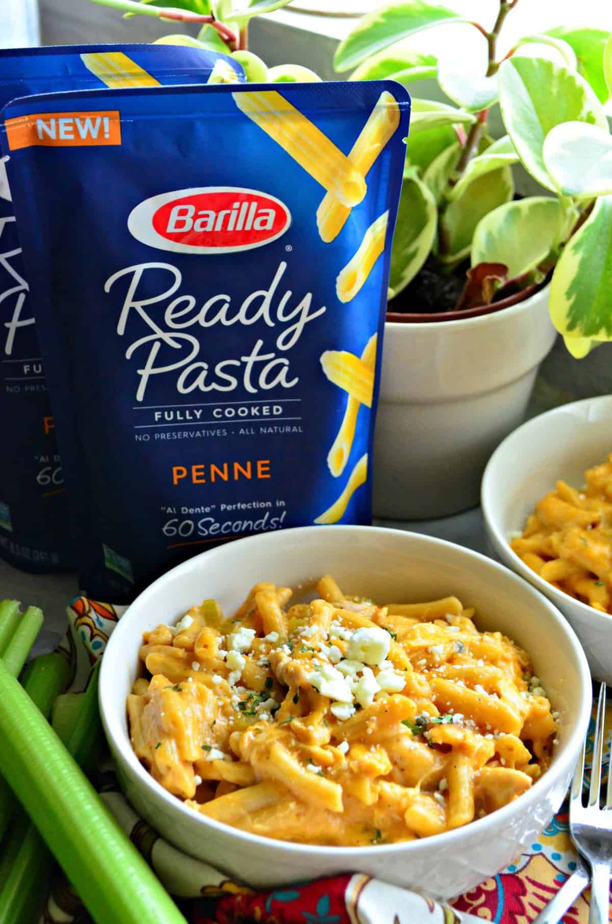 bowl of creamy buffalo chicken pasta next to celery and in front of 2 bags of barilla ready penne pasta.