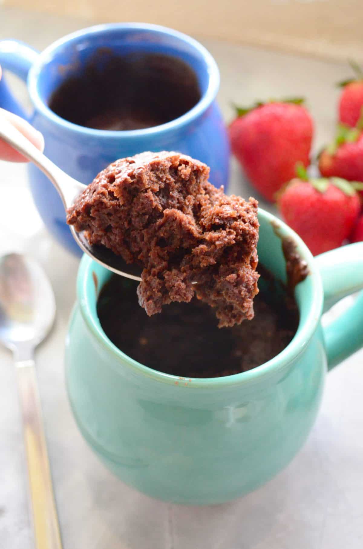 spoonful of chocolate cake held over 2 mugs of cake with strawberries on countertop by it.