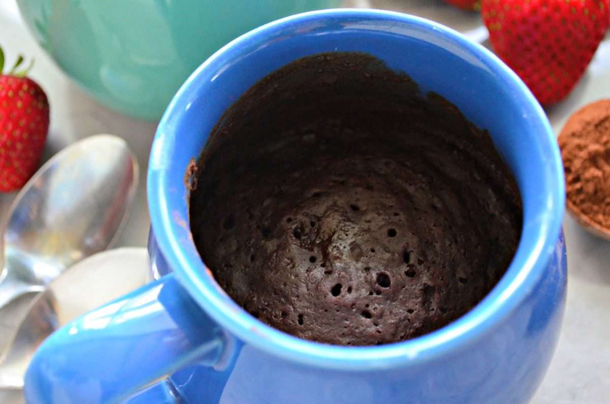 closeup of blue mug filled with brown cake with strawberries blurred in background.