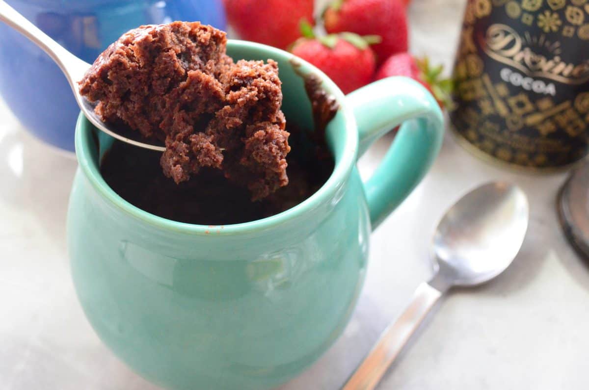 spoonful of chocolate cake held over turquoise mug with strawberries and spoon in background.