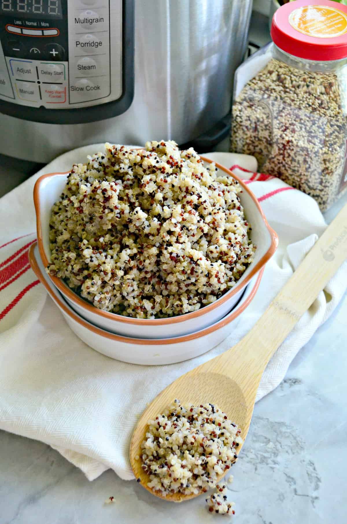 multi-colored cooked quinoa in white bowl with orange rim on cloth with wooden spoon in front of instant pot.