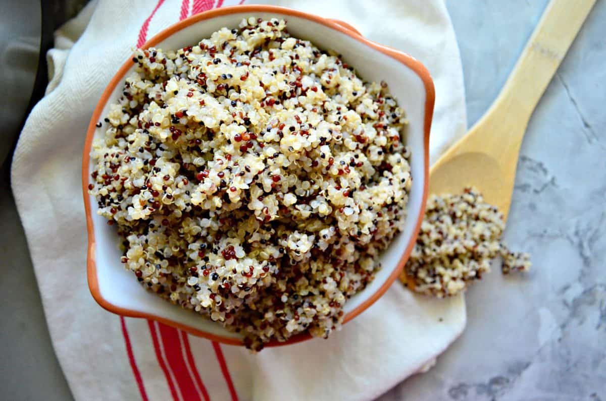 top view of multi-colored cooked quinoa in white bowl with orange rim on cloth with wooden spoon.