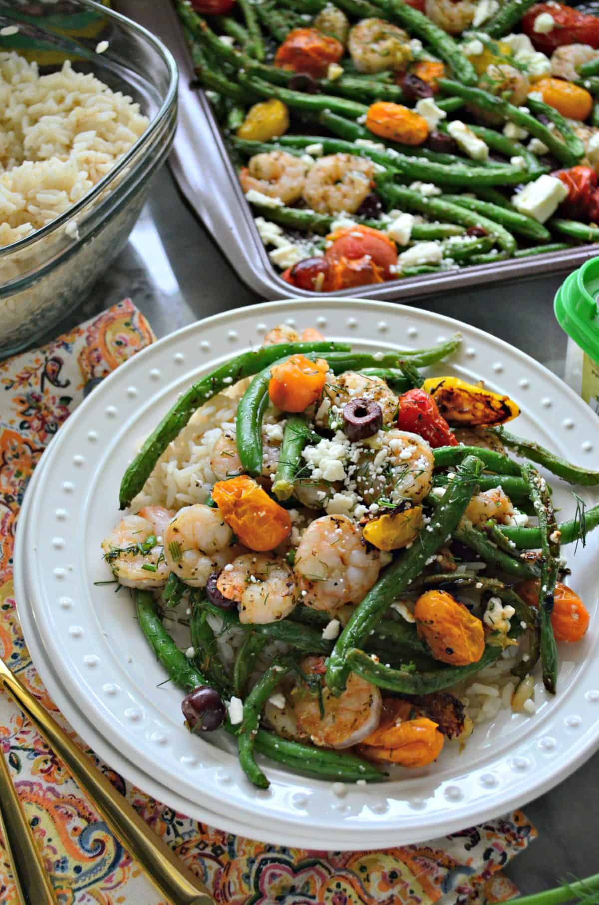 plated feta, green beans, cherry tomatoes, kalamata olives, and dill next to rice dish and sheet pan.