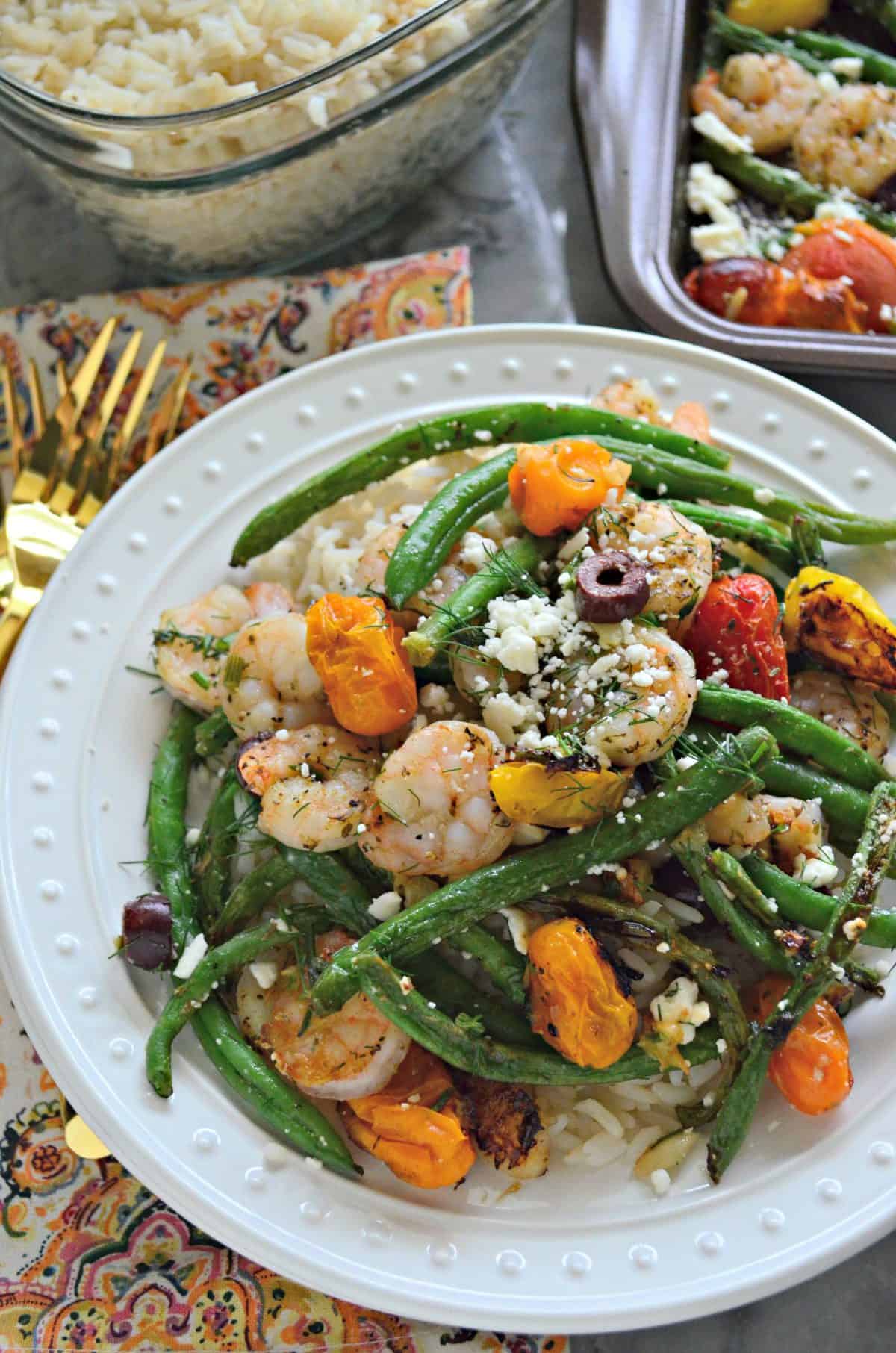 close up plated feta, green beans, cherry tomatoes, kalamata olives, and dill next to rice dish and sheet pan.