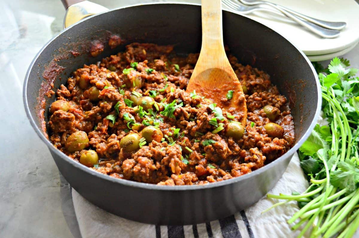 ground meat, red sauce, green olives, and fresh herbs in a skillet with wooden spoon next to fresh cilantro.